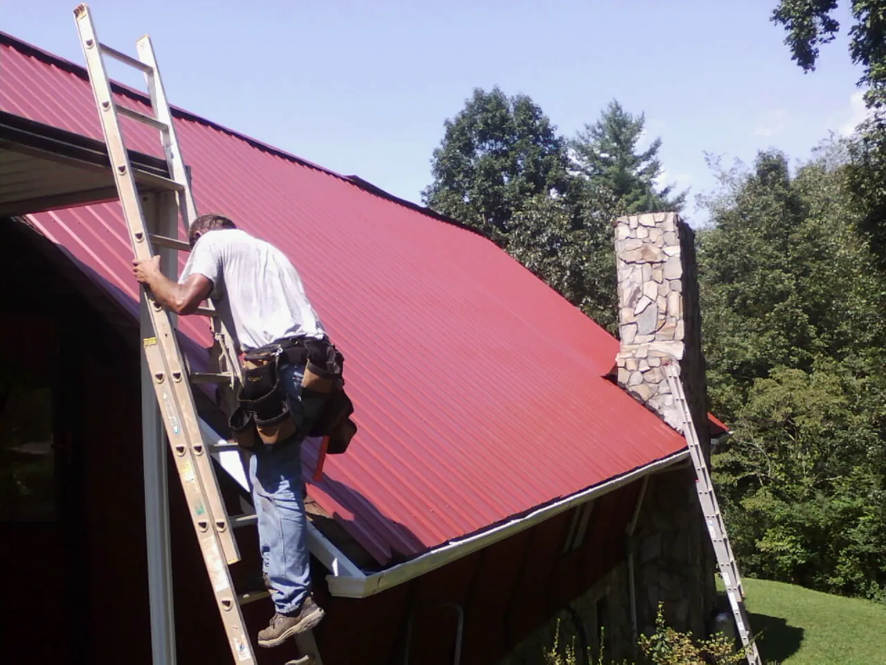 Roofer on a ladder beside a red metal roof and stone chimney — Soffit & Fascia Repair by Roofing Brothers in Mosheim